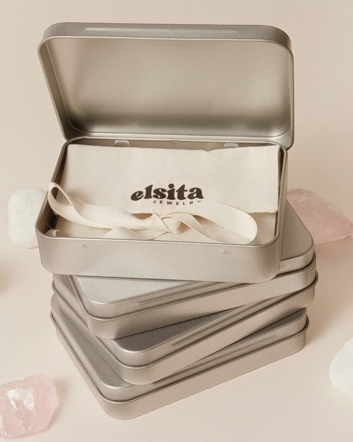 Elsita Jewelry branded metal gift tin containing a fabric pouch, surrounded by raw stones on a neutral background.