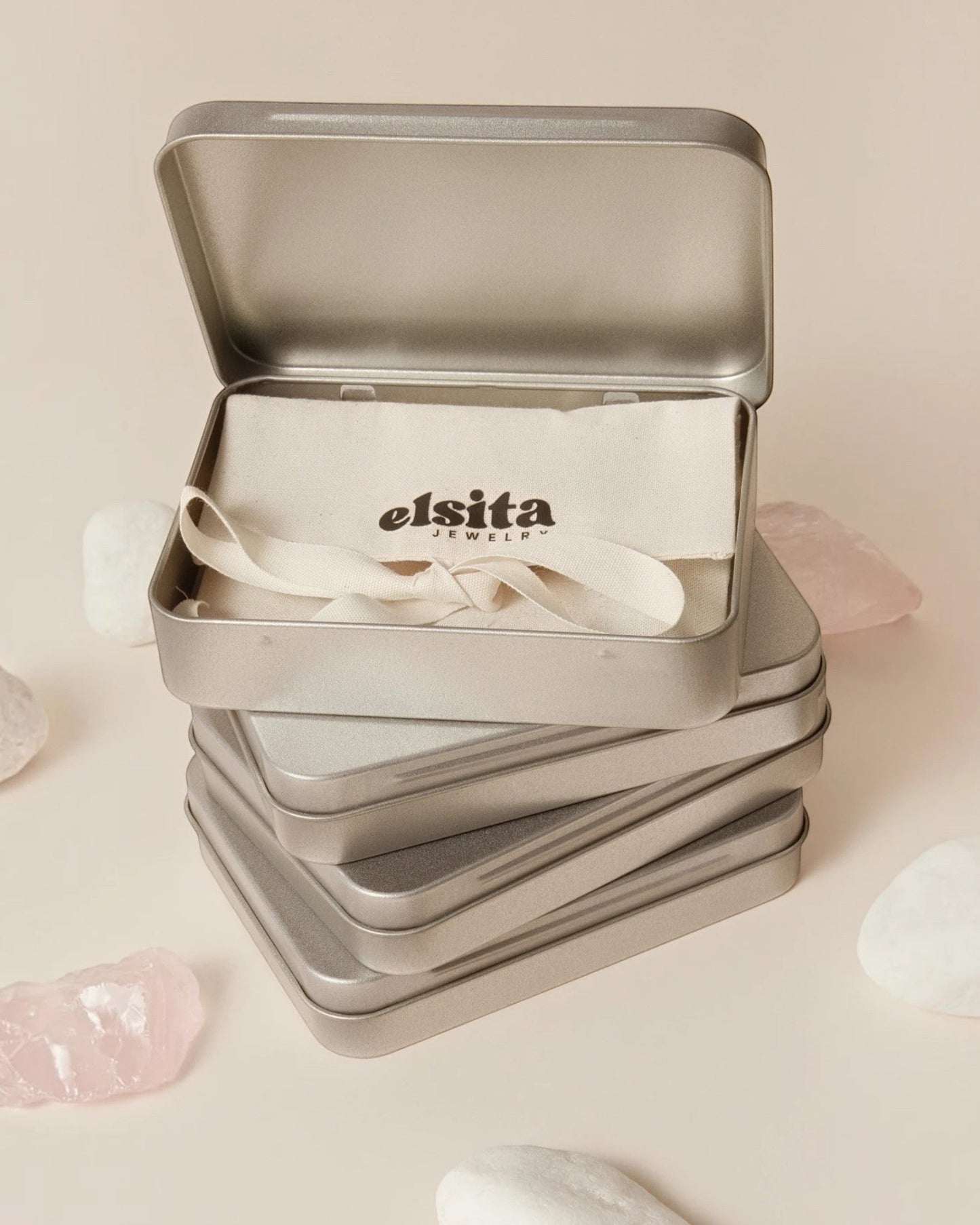 Elsita Jewelry branded metal gift tin containing a fabric pouch, displayed on a beige surface with raw stone accents.