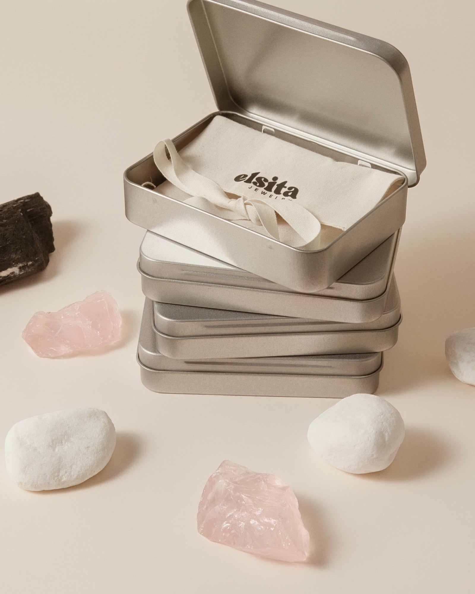 Elsita Jewelry branded metal tins containing Wild 1 agate earrings, displayed with raw rose quartz and white stones.