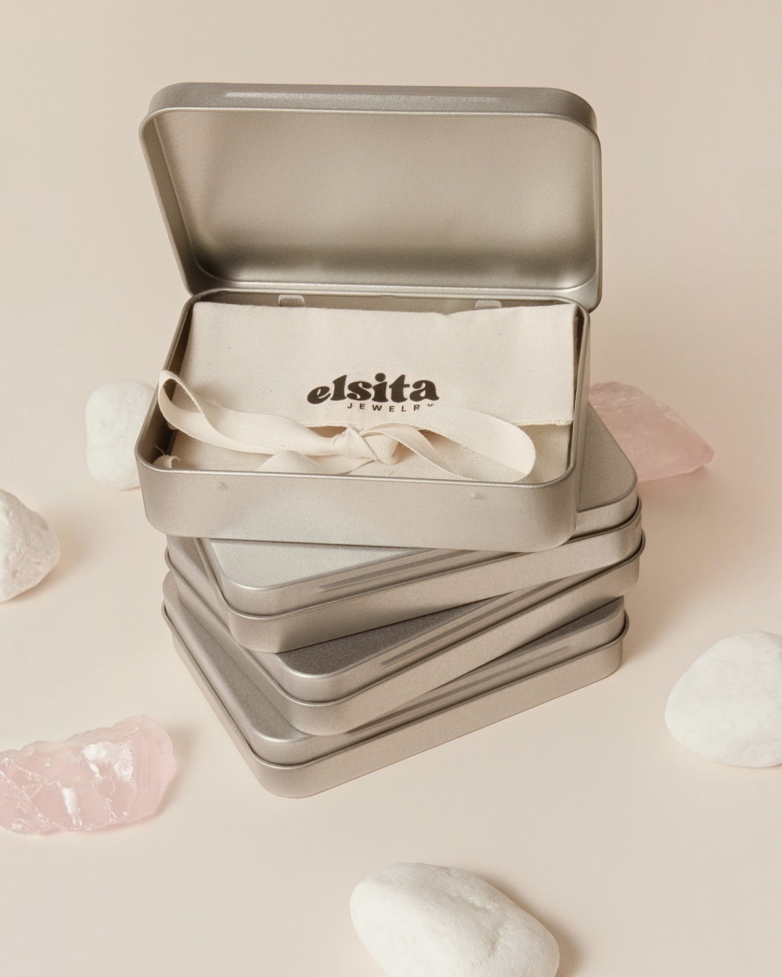 Elsita Jewelry branded metal gift box containing a fabric pouch, displayed with raw mineral stones on a beige surface.