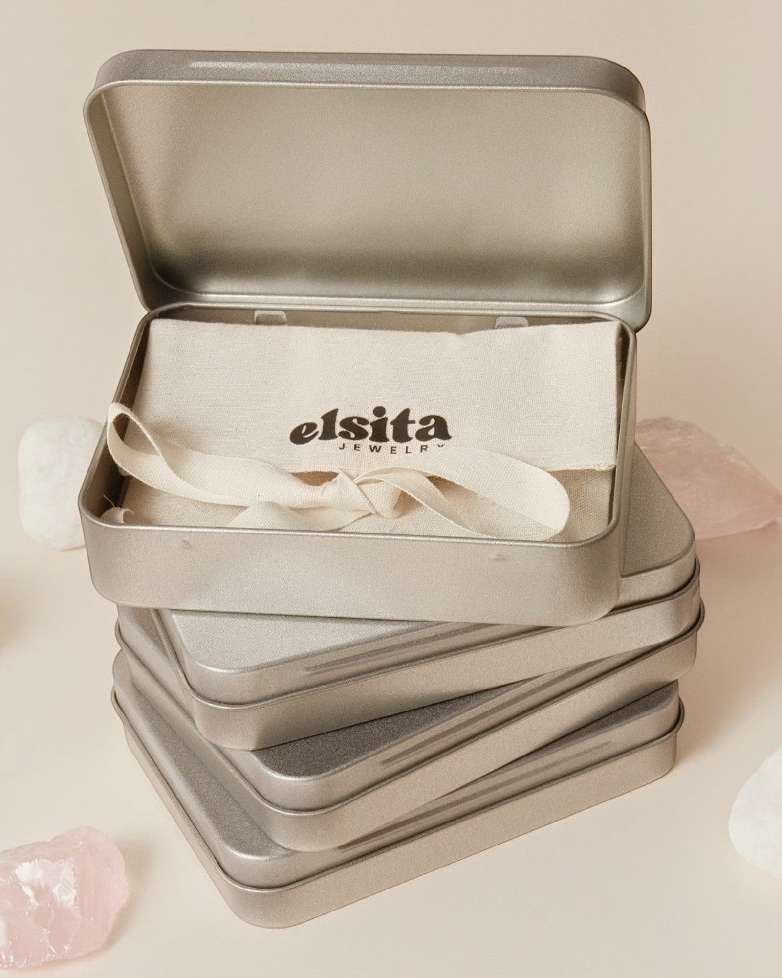 Elsita Jewelry branded metal gift tin containing a fabric pouch, surrounded by raw stones on a neutral background.