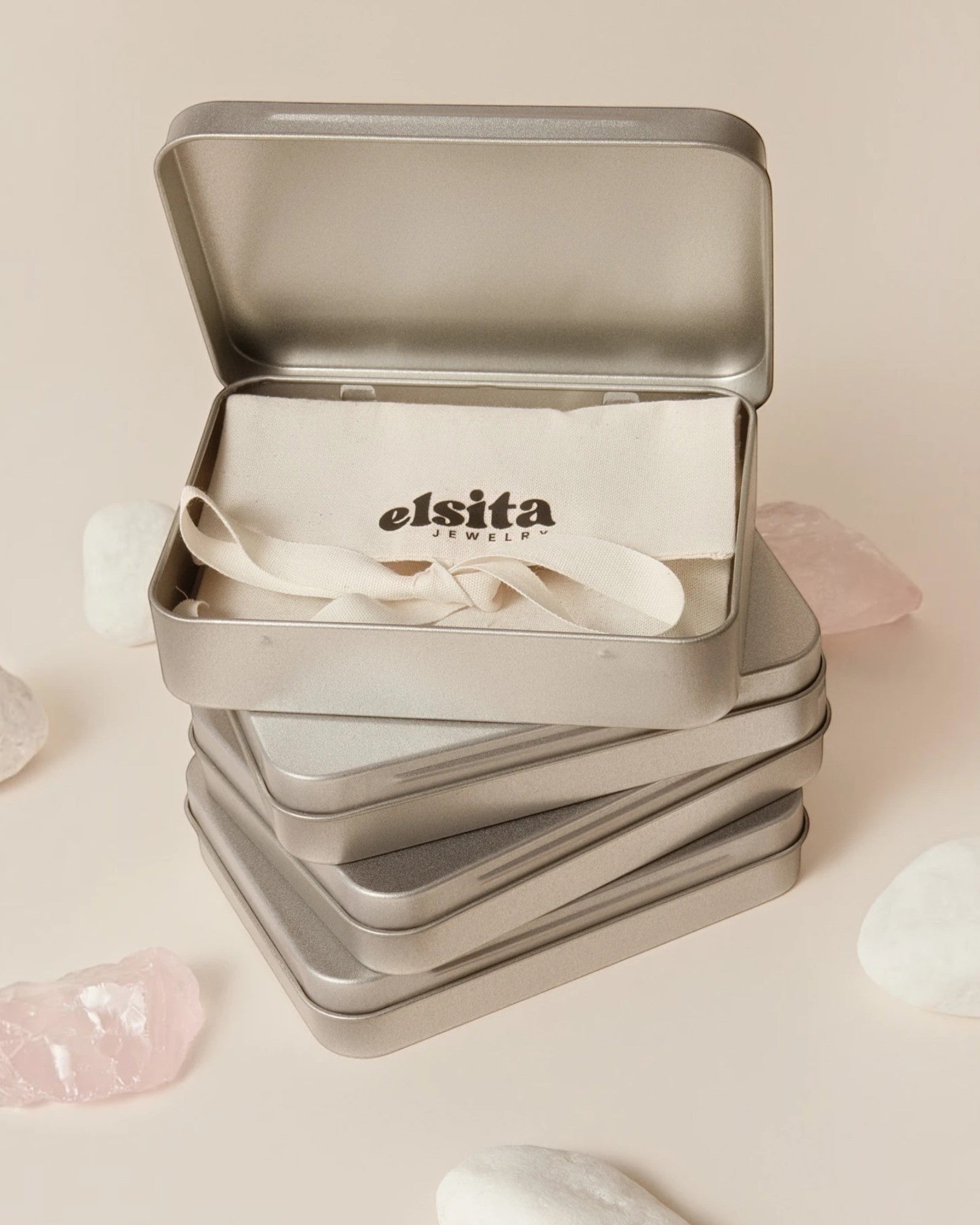 Elsita Jewelry branded metal gift tin containing a fabric pouch, displayed on a beige surface with raw stone accents.