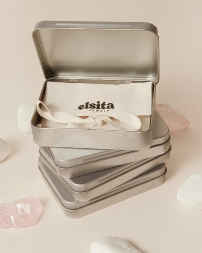 Elsita Jewelry branded metal gift tin containing a fabric pouch, displayed on a beige surface with raw stone accents.