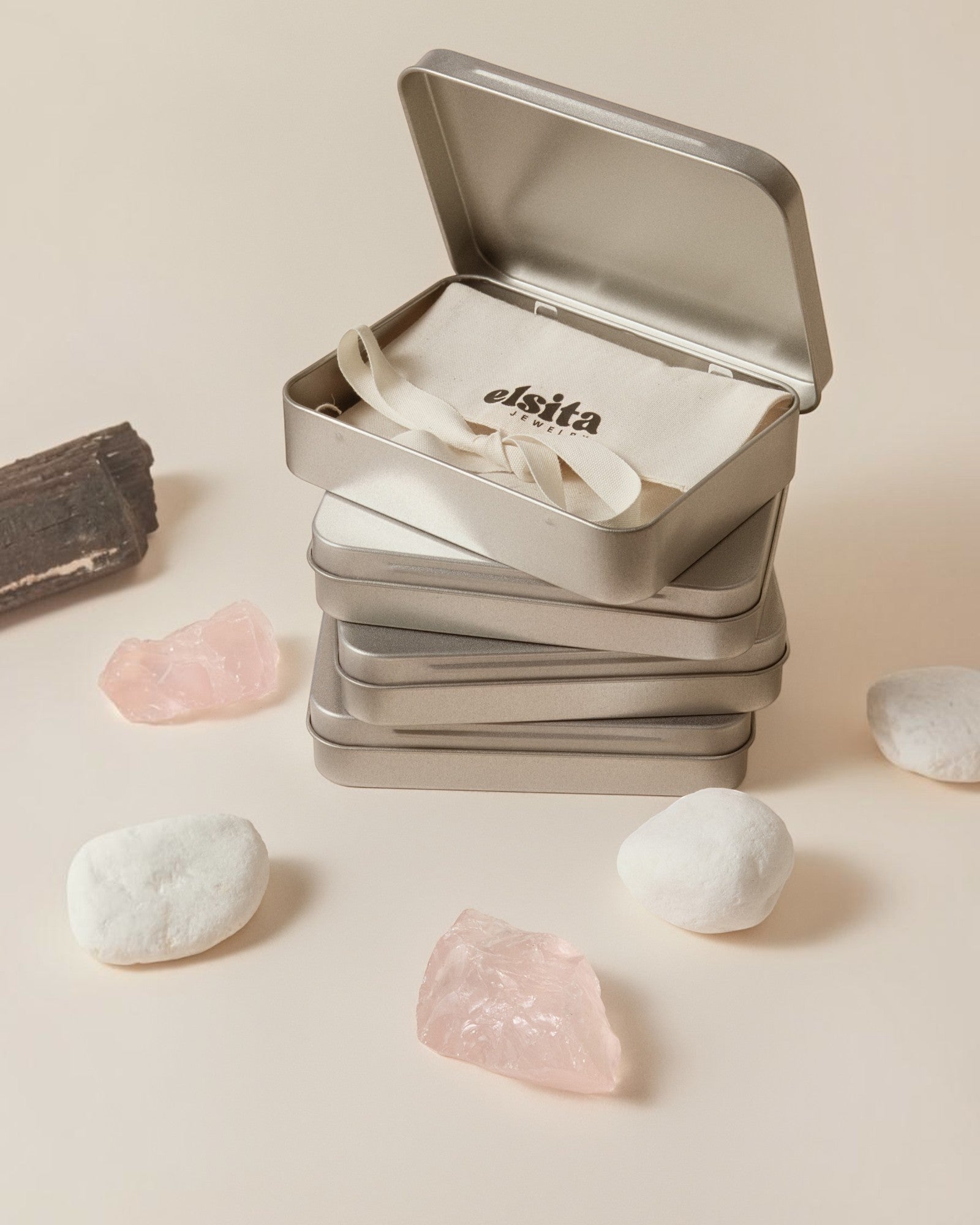 Elsita Jewelry branded metal gift tins stacked with a fabric pouch, surrounded by raw rose quartz and white stones.