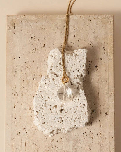 Elsita Jewelry Lustre II clear quartz pendant necklace on a gold chain resting on a textured stone block.