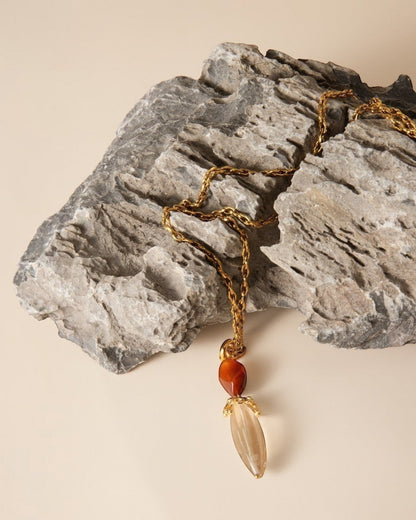 Elsita Jewelry Flame necklace featuring a clear quartz pendant and red agate on a textured gold chain resting on rock